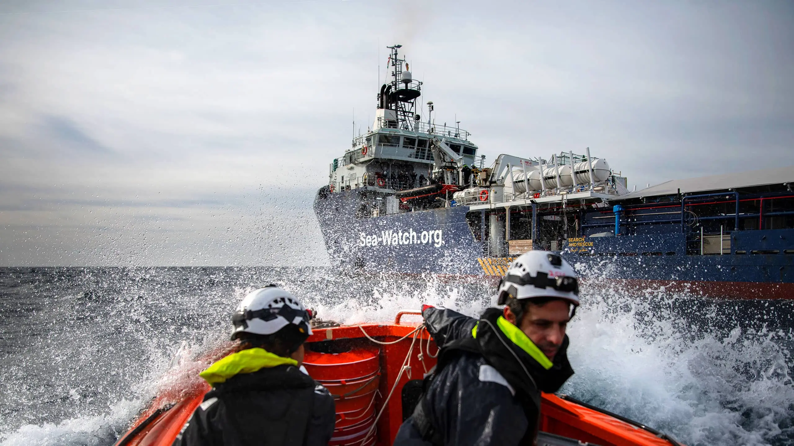 Ein Rettungsteam in einem kleinen Schlauchboot fährt durch aufspritzende Wellen auf das Sea-Watch-Rettungsschiff zu, das im Hintergrund sichtbar ist.